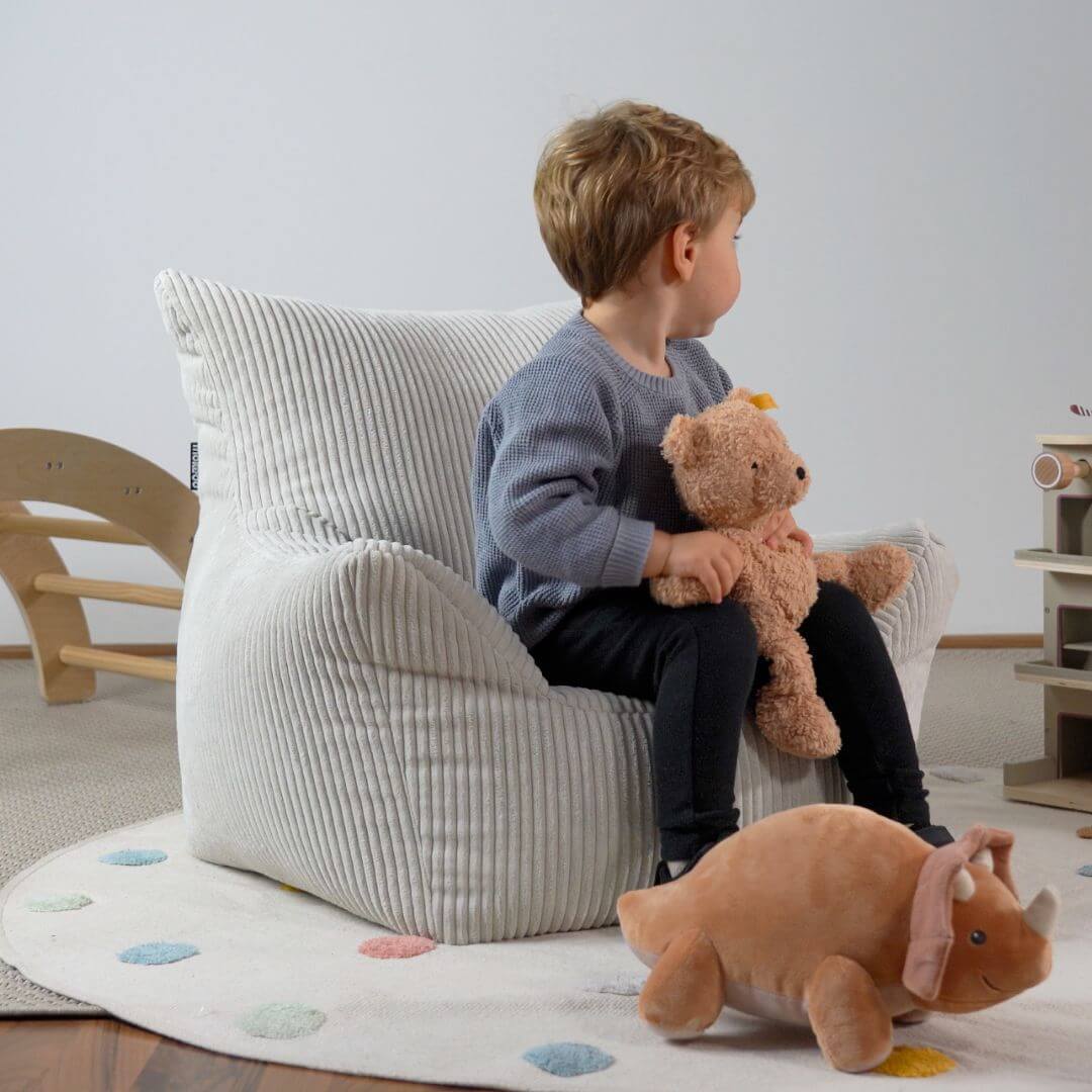 Kindersitzsack aus Cord in beige im Kinderzimmer. Kind sitzt auf Sitzsack, schaut von der Kamera weg und hält einen braunen Teddybär in der Hand 