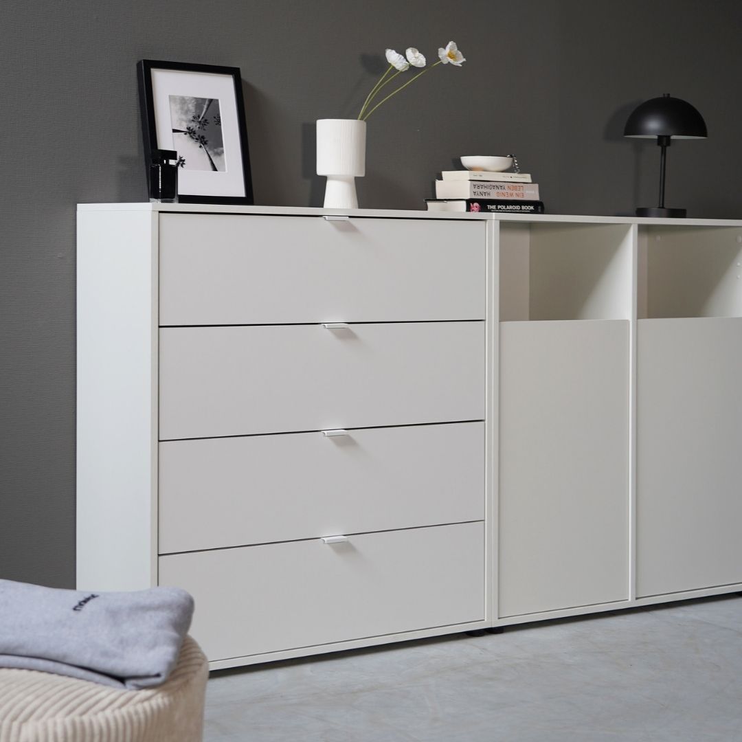 weiße Kommode 'Die Stilvolle' mit vier Fächern neben Wäscheschrank 'Der Wäschehüter' mit zwei Türen an einer grauen Wand 